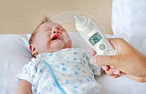 Closeup mother hand takes temperature for infant baby boy with high pressure of ear thermometer on bed at home