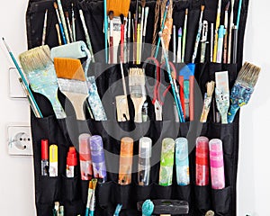 Closeup of colorful dirty paintbrushes, markers and other artistic tools organized in black holder on the white wall