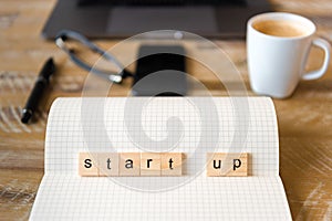 Closeup on notebook over wood table background, focus on wooden blocks with letters making Start Up word