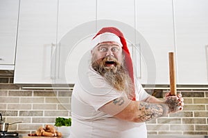 Closeup of an overweigh man in Santa Claus hat with tattooed arms wearing a white T-Shirt, holding a wooden rolling pin on hands