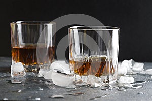 Closeup of pair of ol fashioned glasses.Brandy and pieces of ice on gray table against dark wall