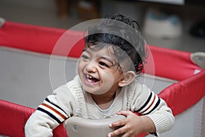 A closeup photo of an adorable indian toddler baby boy smiling with dimple in cheeks and standing inside a playpen