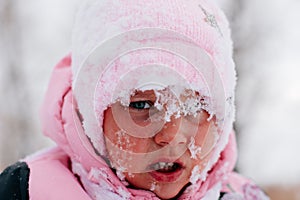 Closeup photo of female kid with sad face covered with snow and about to cry wearing pink winter clothes in forest