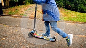Closeup photo of little boy standing and riding on kick scooter at autumn park