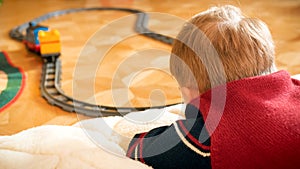 Closeup photo of little boy watching his toy train riding on railways on wooden floor at house