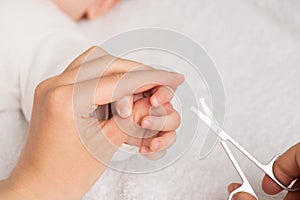 Closeup photo of mother`s hands holding newborn`s fingers and baby nail scissors on isolated white textile background