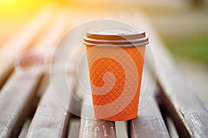 Closeup photo of orange cardboard paper coffee cup standing on the wooden bench in the park with blurred background.