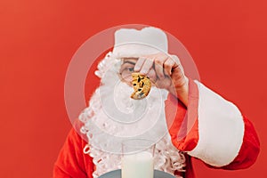 Closeup photo of a Santa`s hand holding a cookie and putting this goodie into a glass of milk. Focus on a cookie, red background