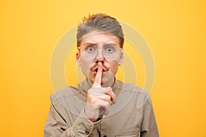 Closeup photo of surprised young man in glasses and mustache on yellow background, shows silence sign and with shocked face looks