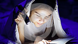 Closeup photo of teenage girl reading book with under blanket with flashlight