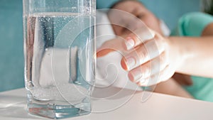 Closeup photo of young sick woman reaching for glass of water with aspirin on bedside table