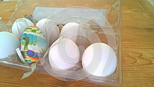 A closeup photograph of a single painted plastic Easter egg nested inside of a plastic egg carton with several real chicken eggs