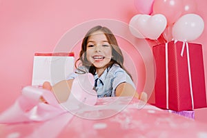 Closeup pink present giving little joyful girl to camera on pink background. Smiling suround big giftboxes, balloons