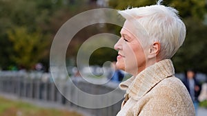 Closeup portrait 65s happy smiling mature lady in casual clothes standing posing outdoors turns head looking aside. Calm