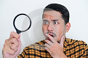 Closeup portrait of adult Asian man showing shocked expression when looking through magnifying glass