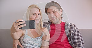 Closeup portrait of adult attractive caucasian couple taking selfies on phone smiling happily sitting on couch indoors