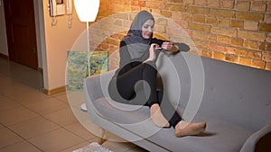 Closeup portrait of adult attractive muslim female in hijab texting on the phone while sitting laidback on the couch