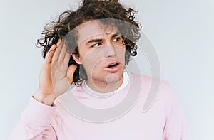 Closeup portrait of amazed eavesdropping male with curly hair placing hand on ear asking someone to speak up, isolated over white
