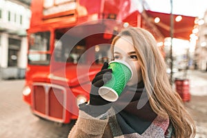 Closeup portrait of attractive girl drinking coffee in the street on red bus background on a cool winter day. Cute girl drinking a