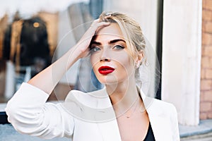 Closeup portrait attractive woman with red lips on street . She wears white jacket, touching hair, looking to camera.