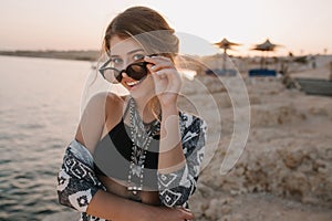 Closeup portrait of attractive young girl taking off stylish sunglasses, on sunset, on beach with sensual look. Wearing