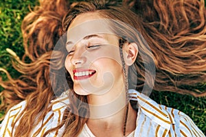 Closeup portrait of a attractive young woman looking directly to the camera lying on the green grass with long red hair outdoors