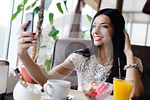 Closeup portrait of beautiful brunette young woman sitting making selfie or selfy on her mobile having fun happy smiling in cafe