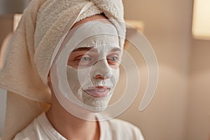 Closeup portrait of beautiful woman with white clay facial mask on face, being wrapped in towel, doing skin care and beauty
