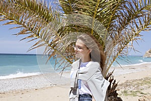 Closeup portrait of a beautiful young Caucasian girl with hair developing from the wind posing near a palm tree on the seashore in