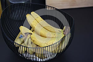 A closeup portrait of a bunch of bananas lying in a decorative black metal bowl on a black kitchen counter. The healthy energizing