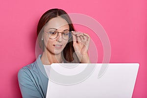 Closeup portrait of charming lady touching frame of her eyeglasses with hand and looking at lap top screen, woman working online,
