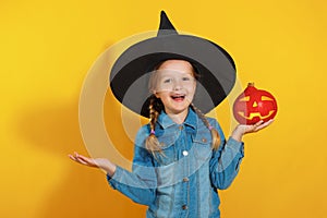 Closeup portrait of a cheerful little girl in a witch hat on a yellow background. Child holds pumpkin jack lantern.