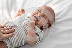 Closeup Portrait Of Crying Little Newborn Baby In Bodysuit Lying On Bed