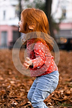 Closeup portrait of cute adorable smiling little red-haired Caucasian girl child playing with dry leaves standing in
