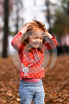 Closeup portrait of cute adorable smiling little red-haired Caucasian girl child playing with dry leaves standing in