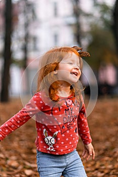 Closeup portrait of cute adorable smiling little red-haired Caucasian girl child playing with dry leaves standing in