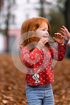 Closeup portrait of cute adorable smiling little red-haired Caucasian girl child playing with dry leaves standing in