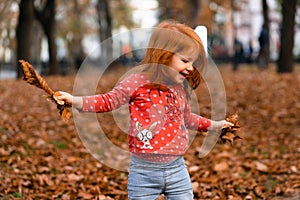 Closeup portrait of cute adorable smiling little red-haired Caucasian girl child playing with dry leaves standing in