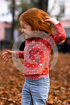 Closeup portrait of cute adorable smiling little red-haired Caucasian girl child playing with dry leaves standing in