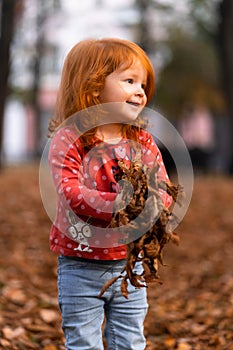 Closeup portrait of cute adorable smiling little red-haired Caucasian girl child playing with dry leaves standing in