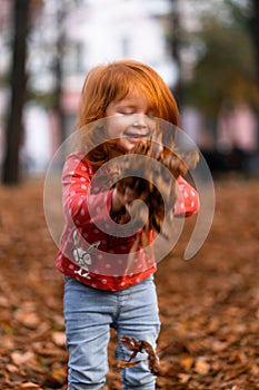 Closeup portrait of cute adorable smiling little red-haired Caucasian girl child playing with dry leaves standing in