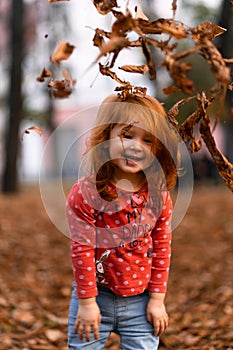 Closeup portrait of cute adorable smiling little red-haired Caucasian girl child playing with dry leaves standing in