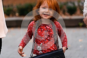 Closeup portrait of cute adorable smiling little red-haired Caucasian girl child standing with mama`s big bag in autumn