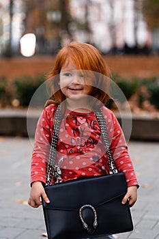 Closeup portrait of cute adorable smiling little red-haired Caucasian girl child standing with mama`s big bag in autumn