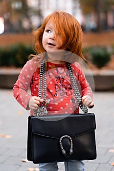 Closeup portrait of cute adorable smiling little red-haired Caucasian girl child standing with mama`s big bag in autumn