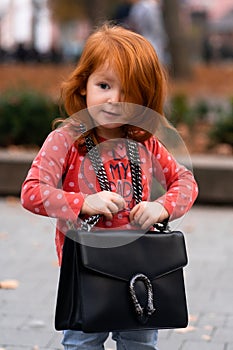 Closeup portrait of cute adorable smiling little red-haired Caucasian girl child standing with mama`s big bag in autumn