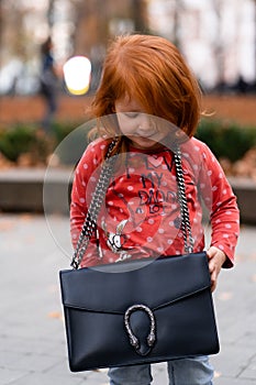 Closeup portrait of cute adorable smiling little red-haired Caucasian girl child standing with mama`s big bag in autumn