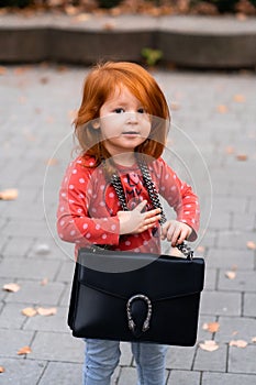 Closeup portrait of cute adorable smiling little red-haired Caucasian girl child standing with mama`s big bag in autumn