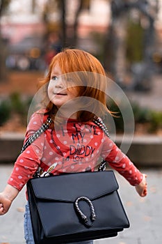 Closeup portrait of cute adorable smiling little red-haired Caucasian girl child standing with mama`s big bag in autumn