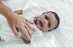 Closeup portrait of cute African american newborn baby lying on bed and looking at camera in bedroom. Mother hand lulling little